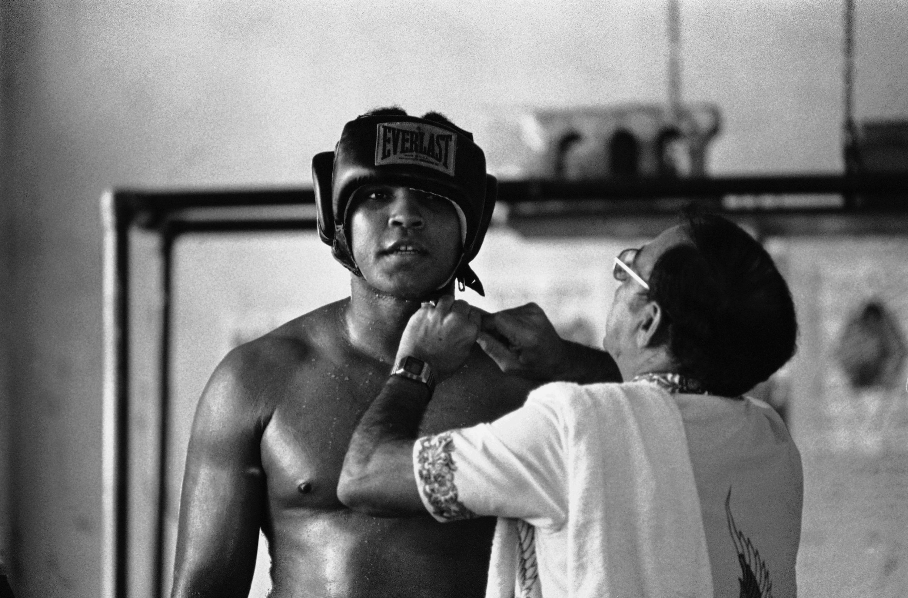 A younger Muhammad Ali being fitted for boxing headgear in a training gym, from the chest up