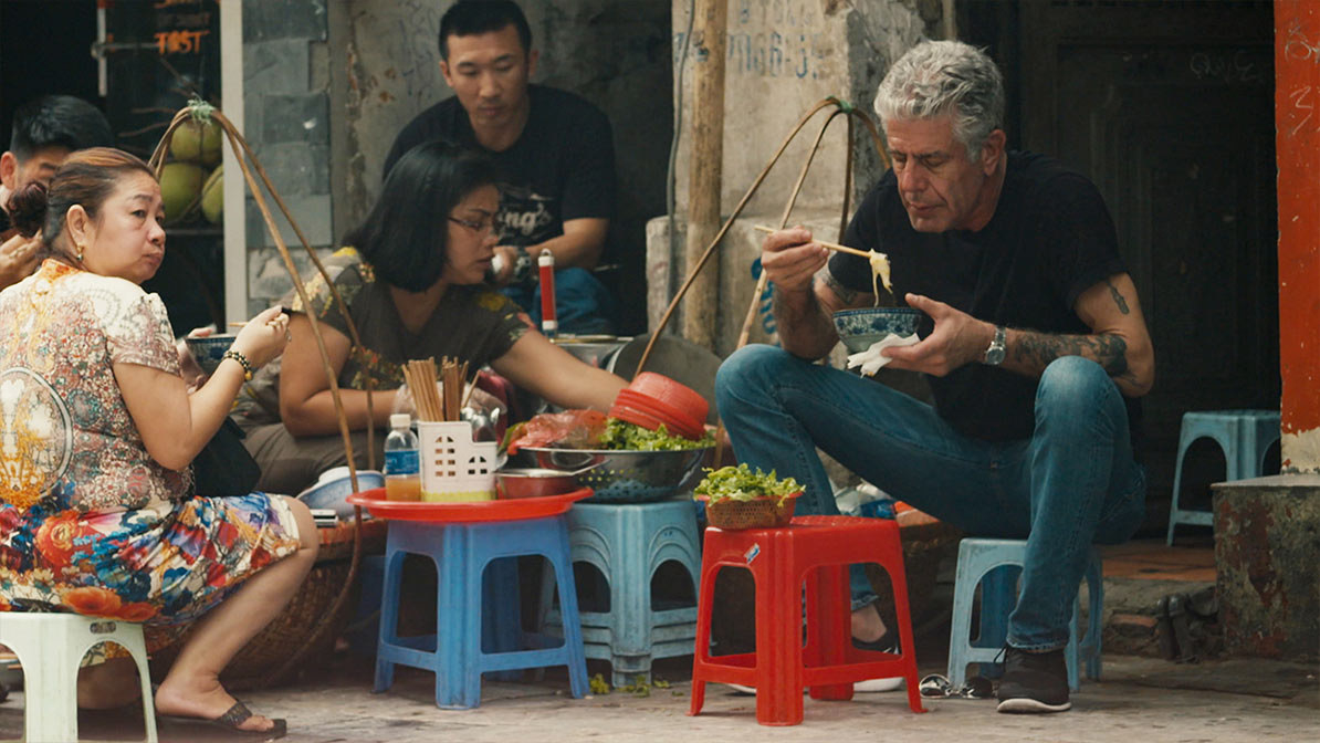 Bourdain about to eat a bowl of noodles, sitting on a small chair in a city street in Asia