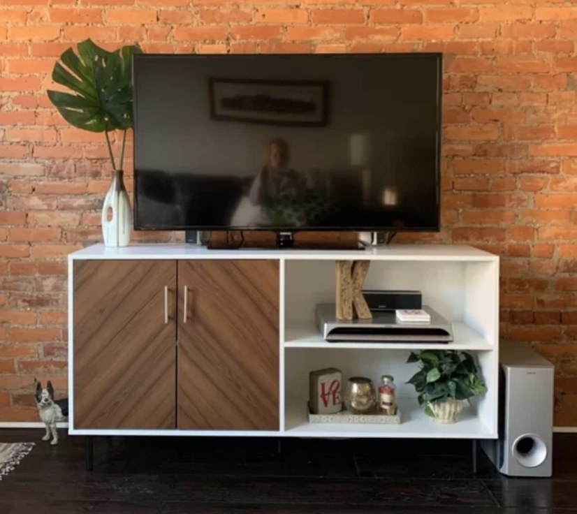 A reviewer&#x27;s acorn/white tv stand with a cabinet and two shelves with a tv atop in a living room