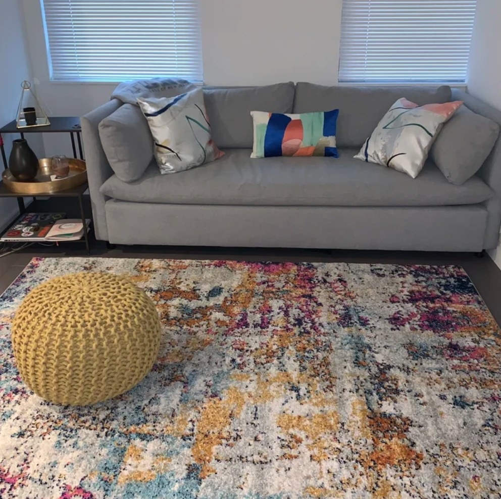 A reviewer&#x27;s yellow/purple/blue, rectangular abstract area rug displayed in a living room