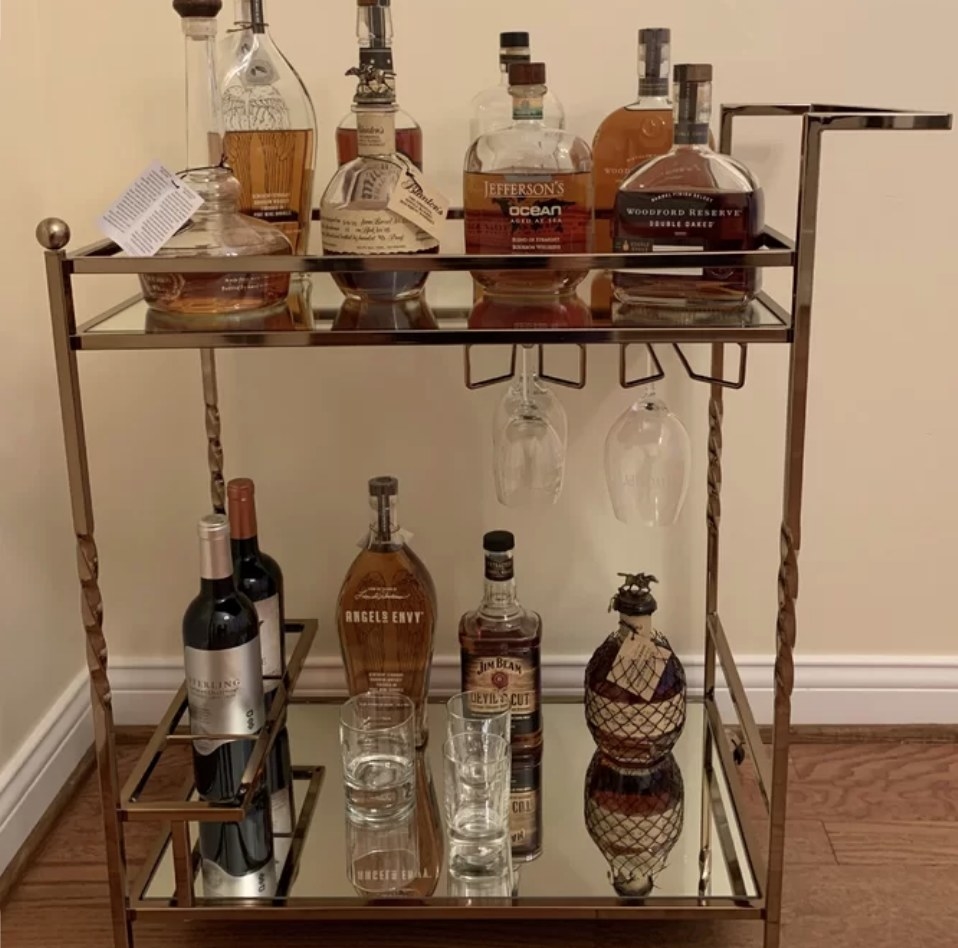 A reviewer&#x27;s champagne and mirrored bar cart with four wheels, two shelves, a wine glass rack stocked with bottles and glasses