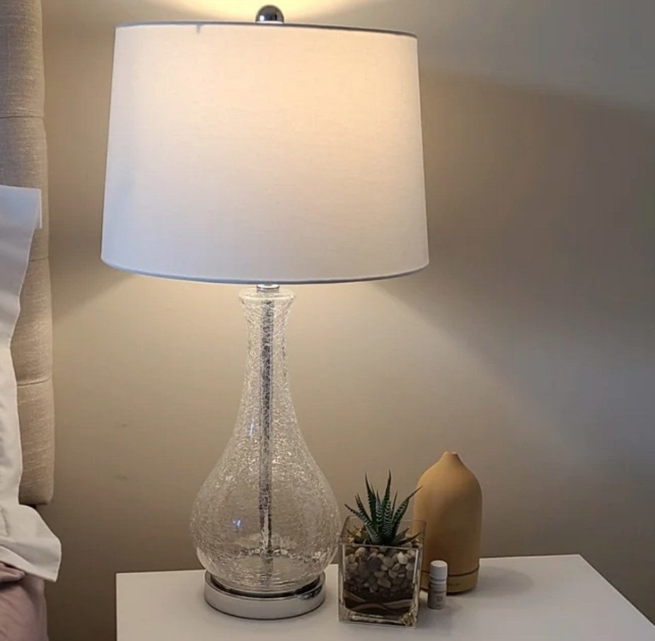 A reviewer&#x27;s clear crackle table lamp with a white dome shade atop a nightstand