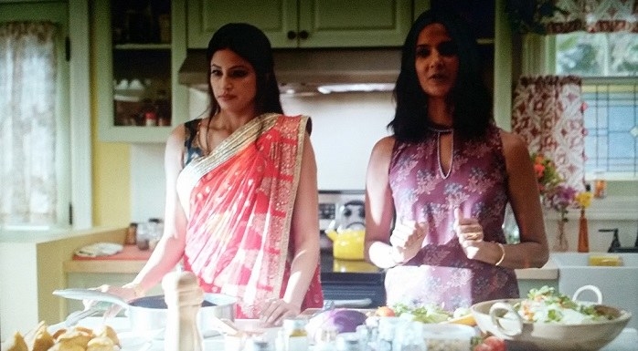 Kamala and Devi&#x27;s mom stand in front of the island in the kitchen. Samosas, a salad, and other foods are on the island. The oven, green cabinets, a white sink, and windows are behind them