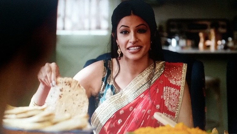 Kamala picks up a roti. She is wearing a red, blue, and gold sari