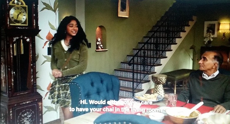 Devi, wearing a white shirt, green cardigan, and green plaid skirt, asks her uncle and Prashant, who are seated at the table, if they want chai in the living room
