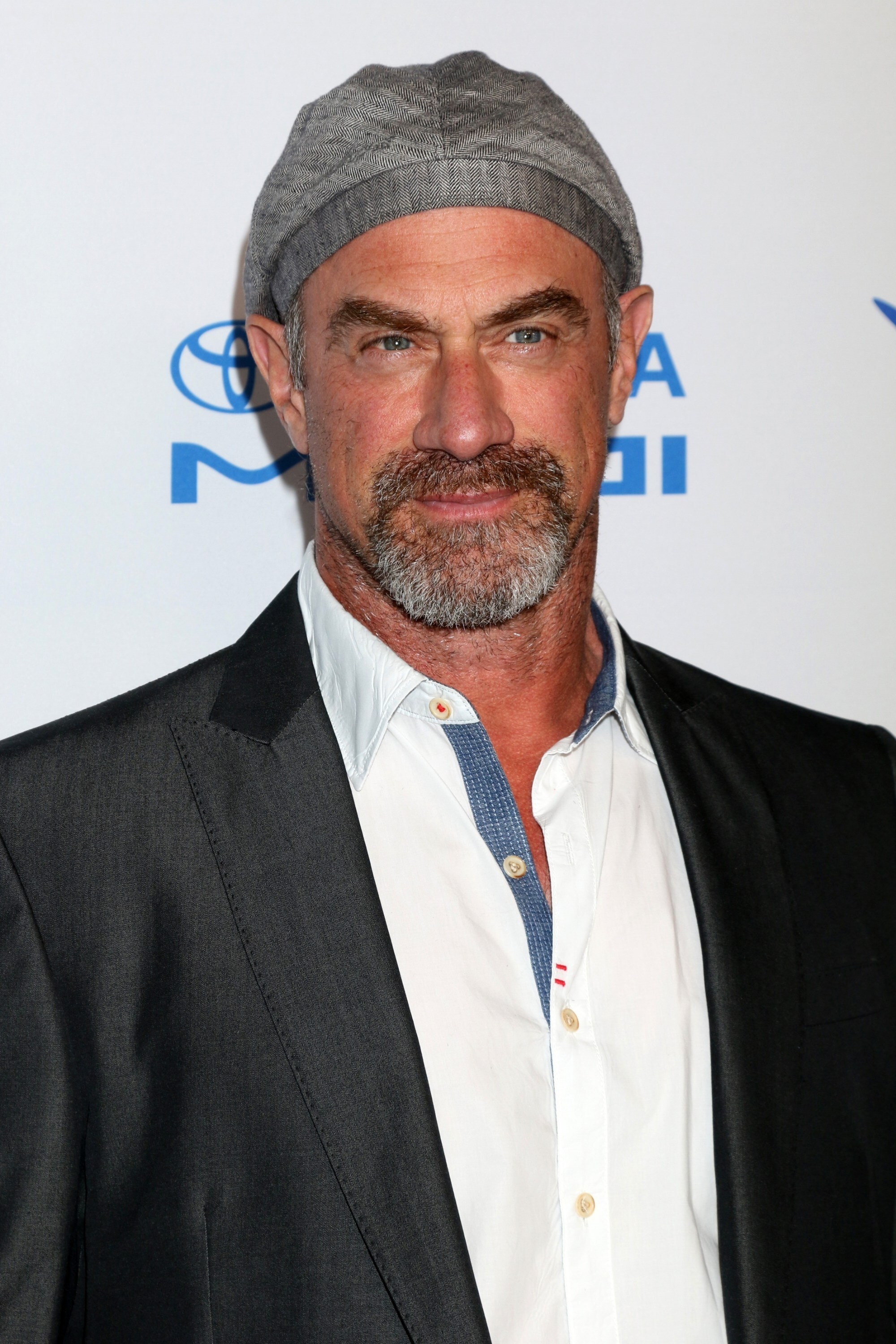 Chest-up portrait of Meloni at a step-and-repeat wearing a button-down and suit jacket with a jaunty beret-style cap, with a subtle half smile