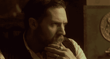tom hardy in peaky blinders in a sweater vest, strokes chin and beard