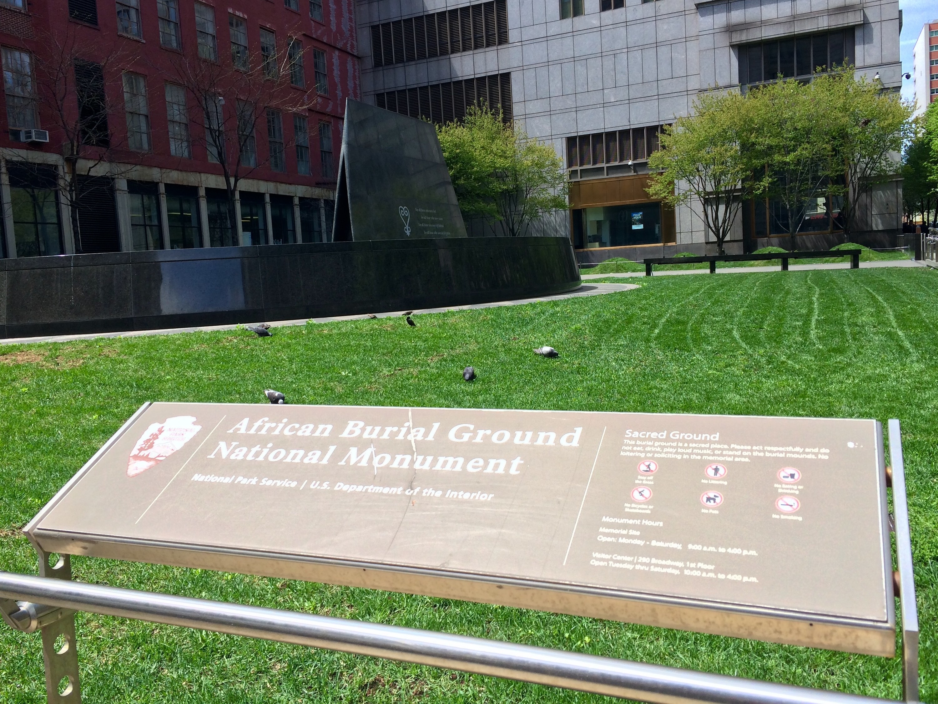 A sign identifying the African Burial Ground national monument hovers in front of an impeccably trimmed grassy area. A dark, marble monument looms over the grass.