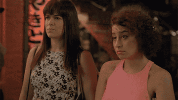 Ilana and Abbi in &quot;Broad City&quot; pushing their mouths up into a smile with their middle fingers