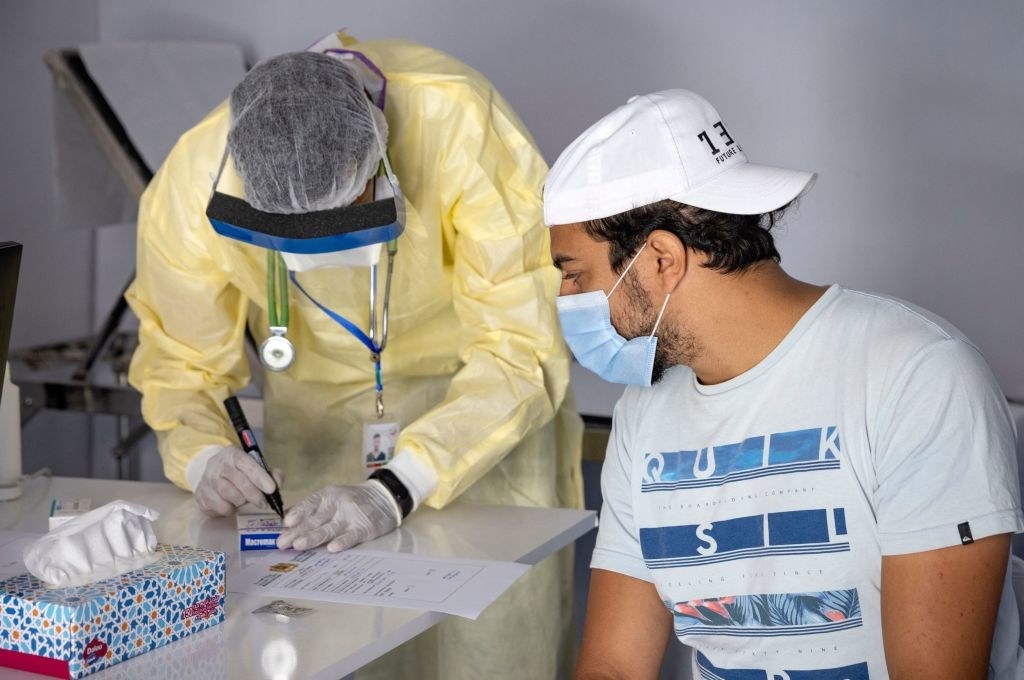 A person in a medical gown and gloves leans over a table while a masked person in a T-shirt and cap watches them