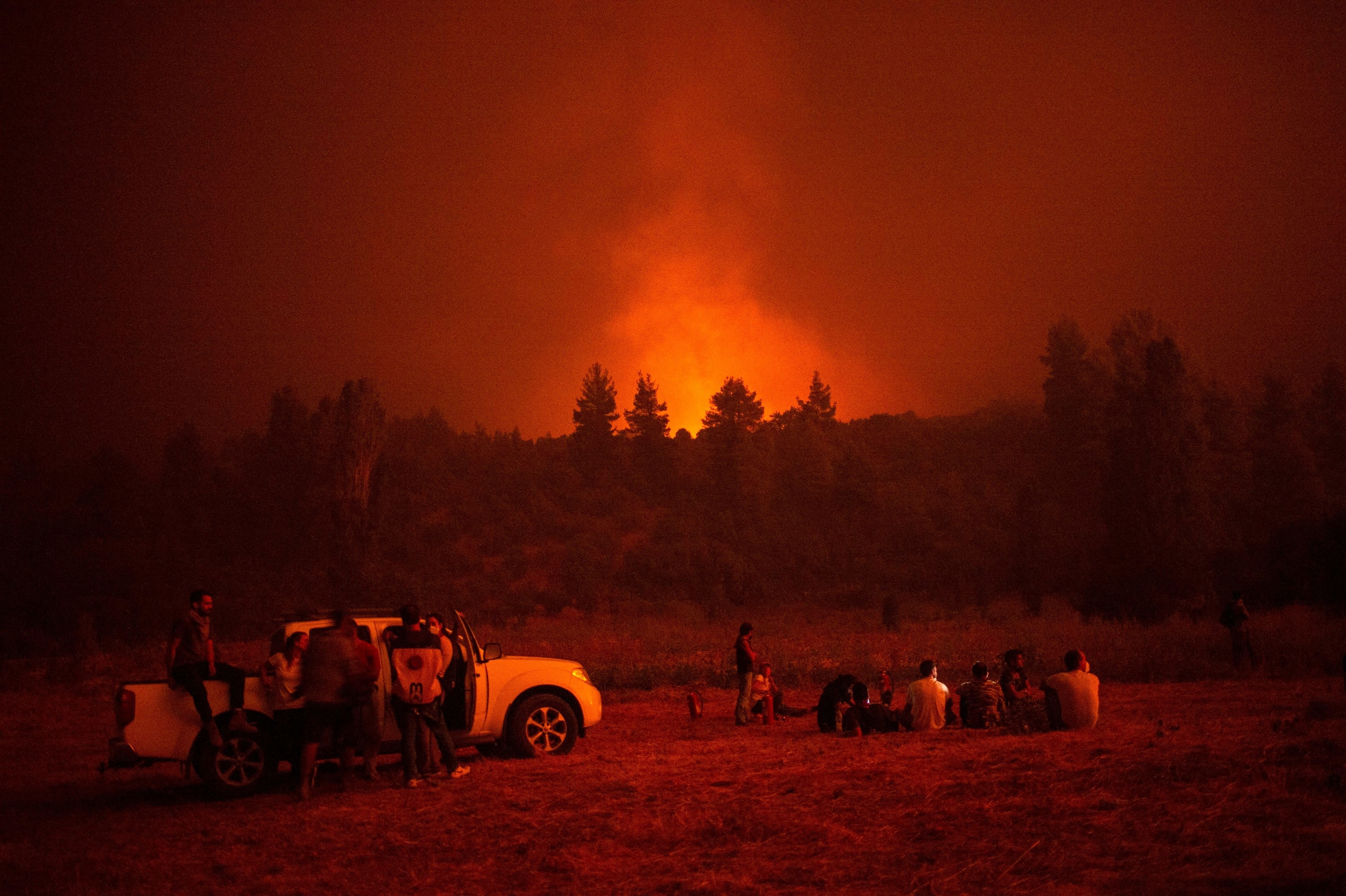 People sit in a meadow under a smoky, hazy sky as a fire burns beyond the horizon