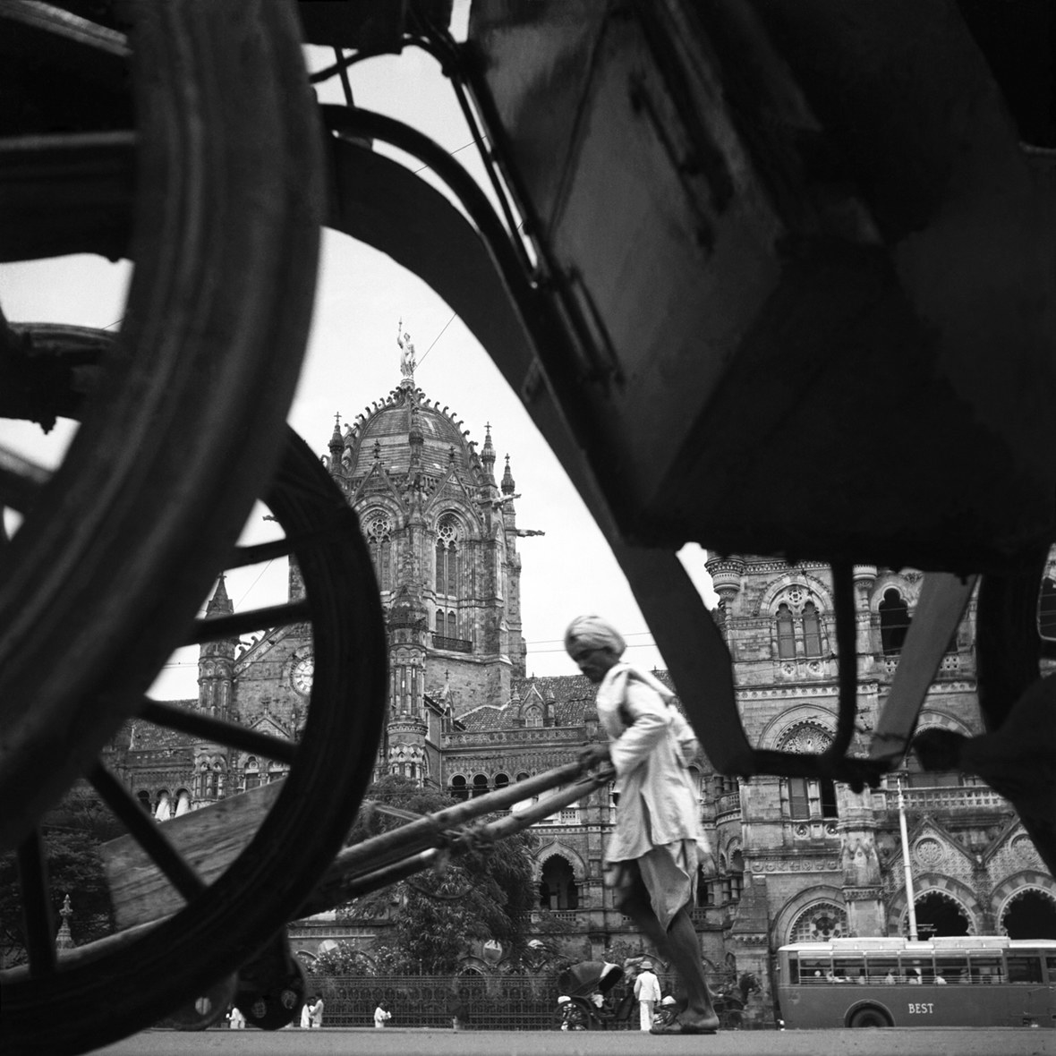 A man pushes a rickshaw in India, seen through the wheels of a cart with an ornate building in the background