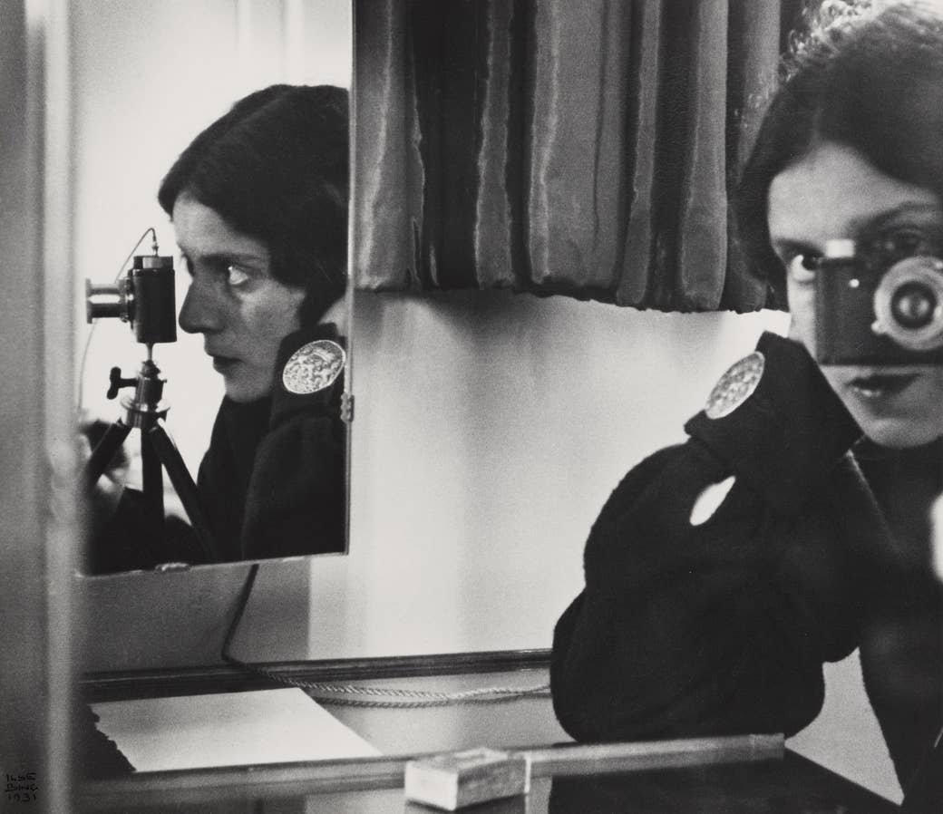 A woman looks through a camera lens while reflected by a mirror on her right