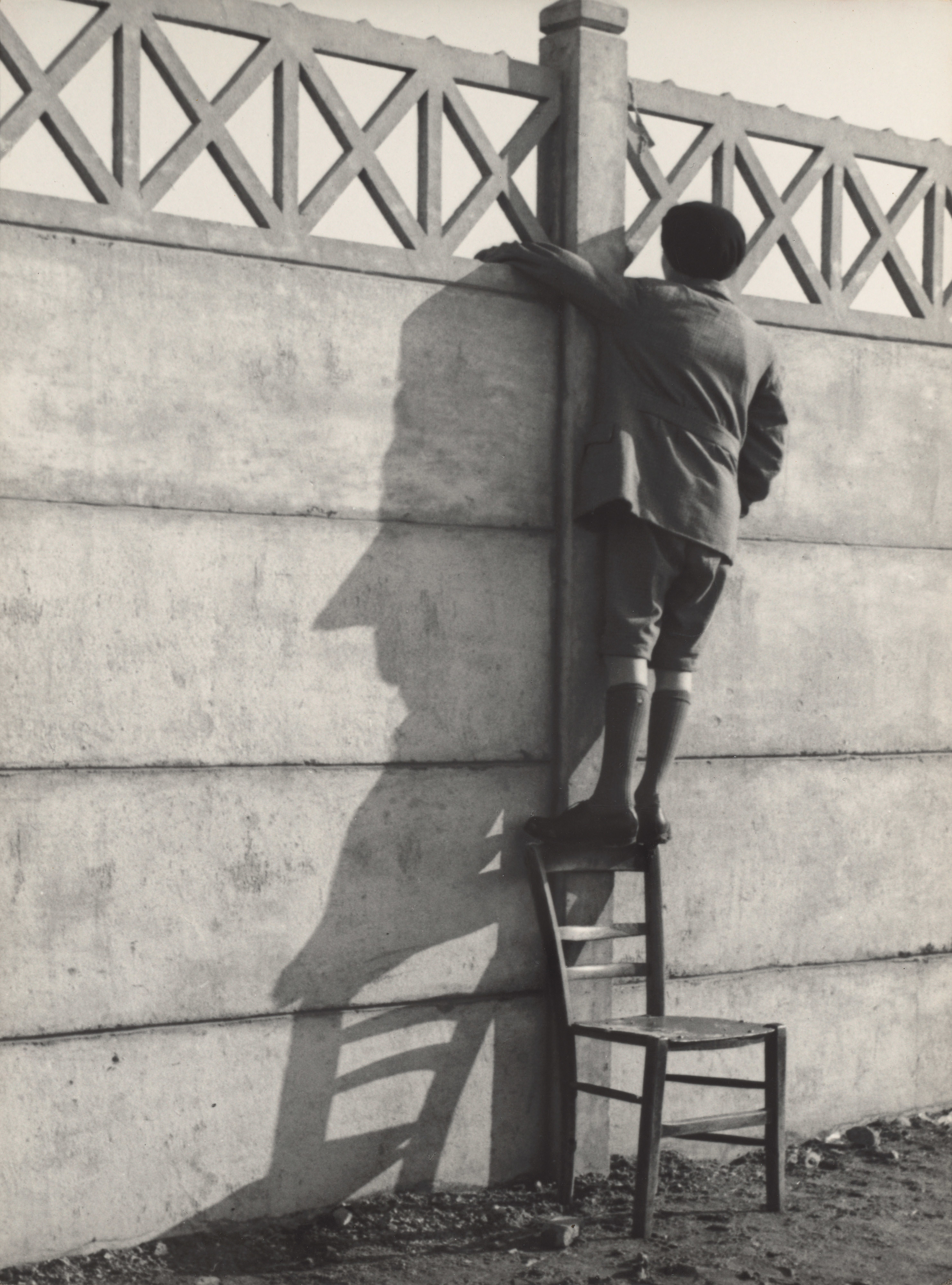 A boy stands on the back of a chair to get a vantage point over a large stone wall