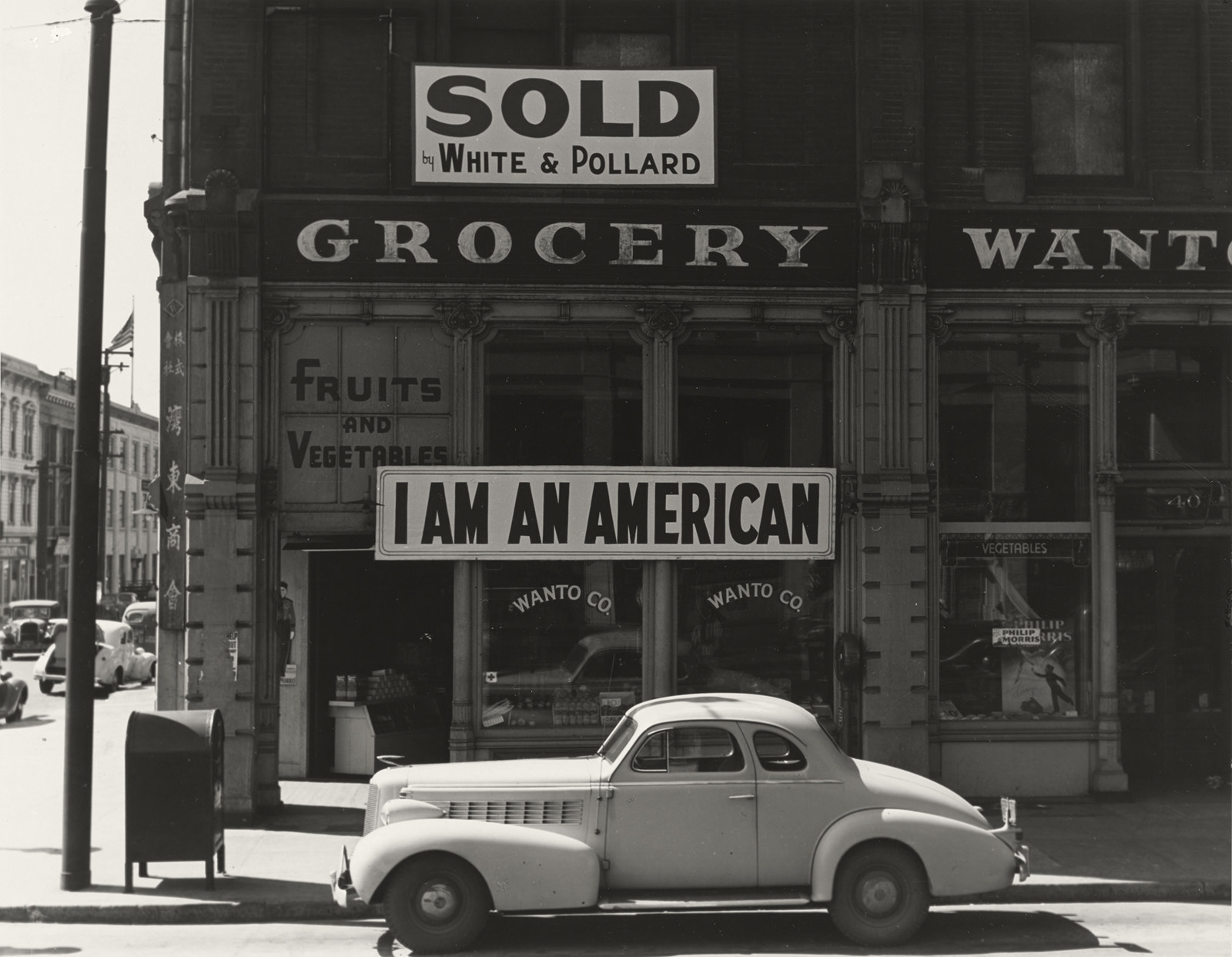 A sign reads I Am An American over a fruit and vegetable store with a car on the street outside