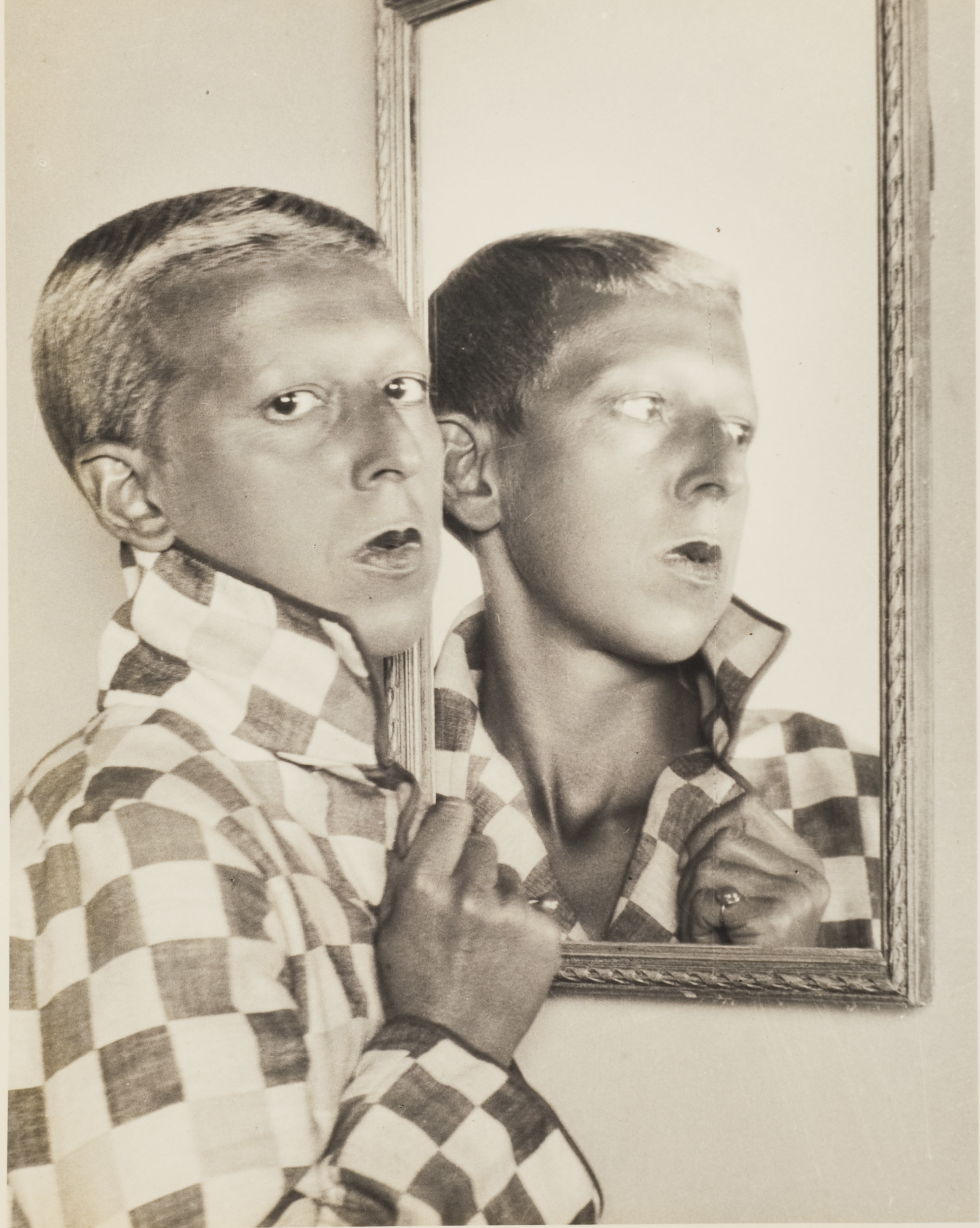 A person with short hair in a checkered jacket, looking back at the camera reflected in a mirror 