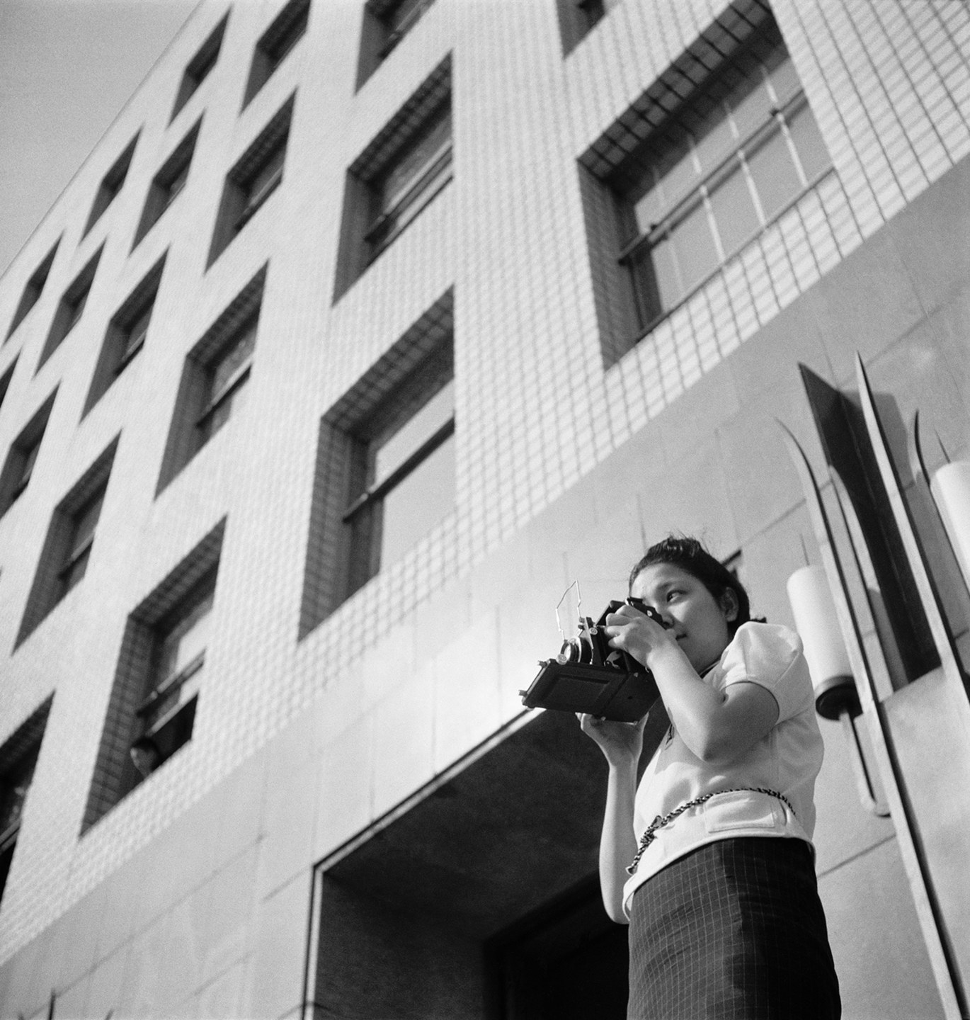 A woman aims a camera while she stands in front of a building