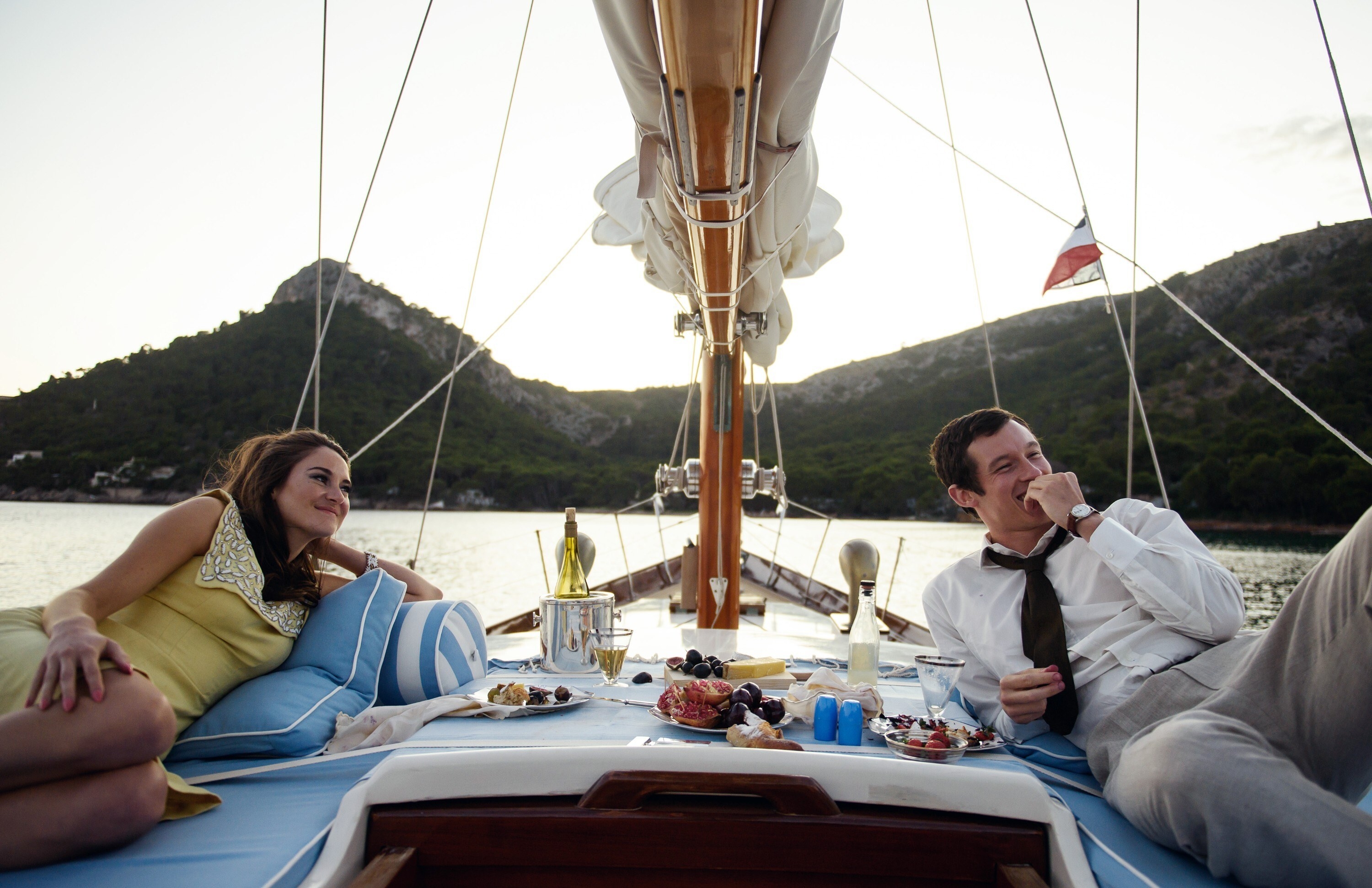 Jennifer and Anthony lounging at the bow of a sailboat with berries and snacks and chilled champagne between them, on a body of water with tree-topped mountains behind them