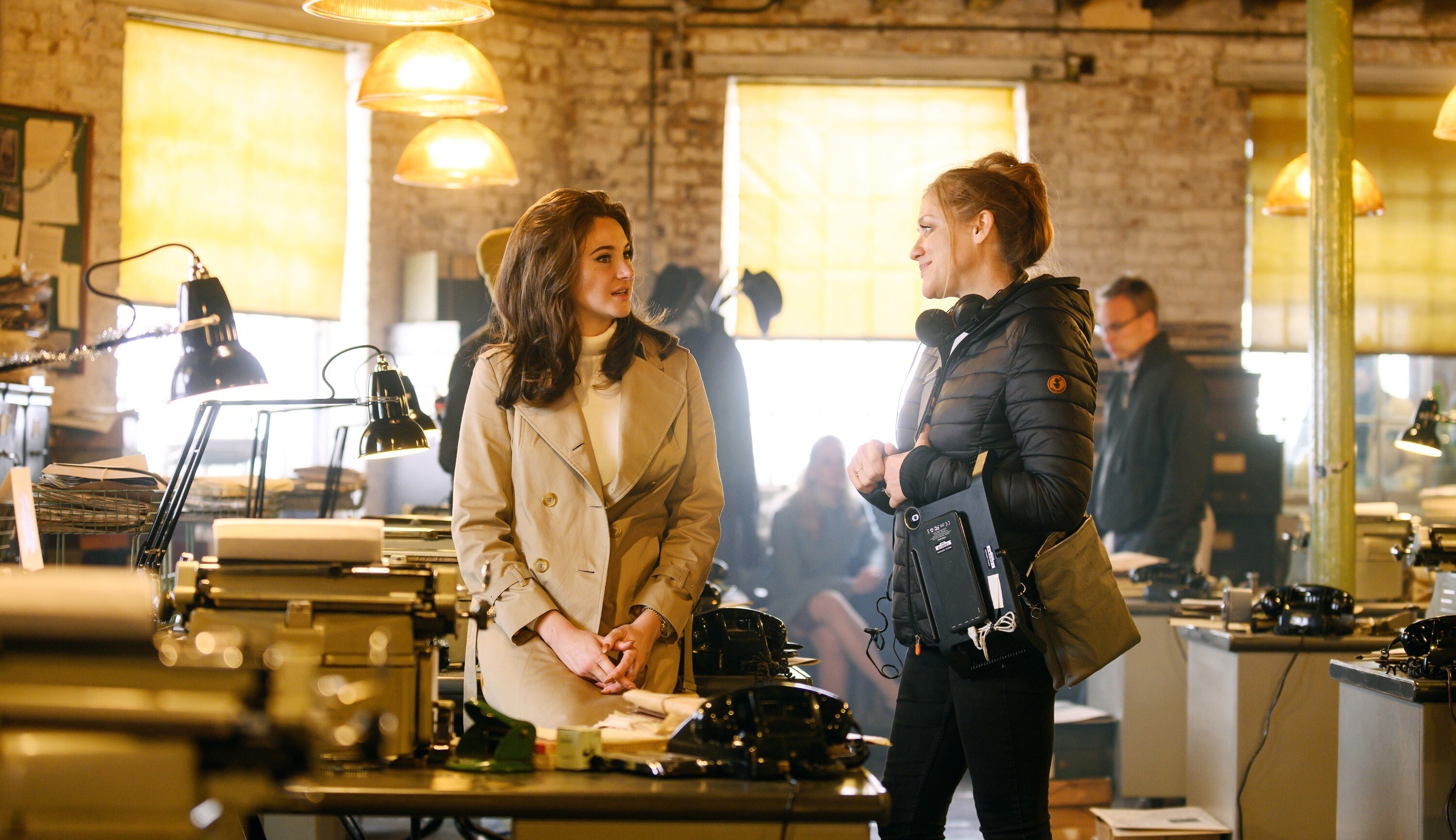 Jennifer in turtleneck and trench coat at the desk of a 1960s office, director Augustine Frizzell in a puffy coat and pants next to her