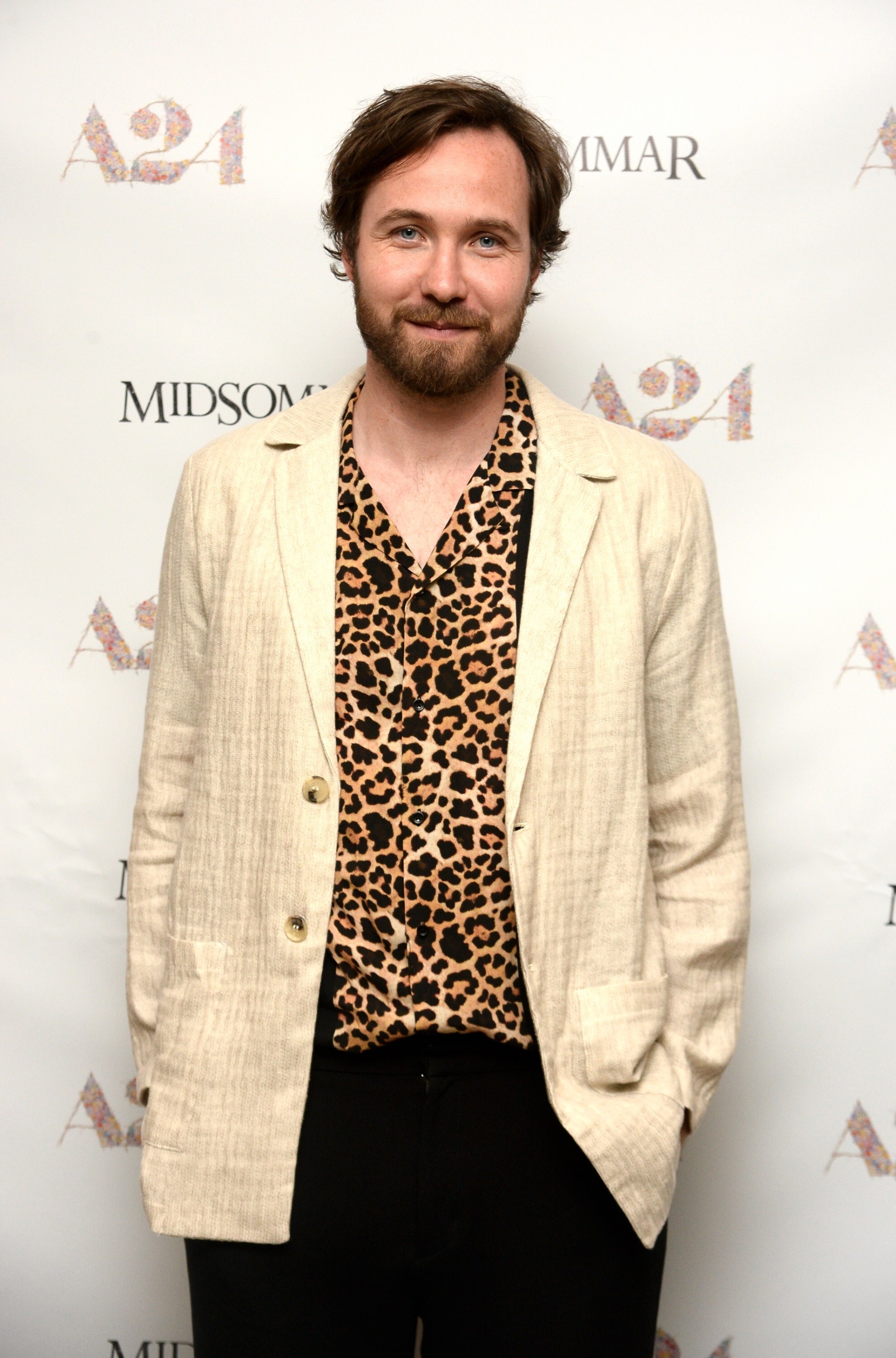 Vilhelm Blomgren in leopard-print button-down shirt and light-colored linen jacket and dark pants standing at a step-and-repeat for a viewing of A24&#x27;s Midsommar, lightly smiling at camera