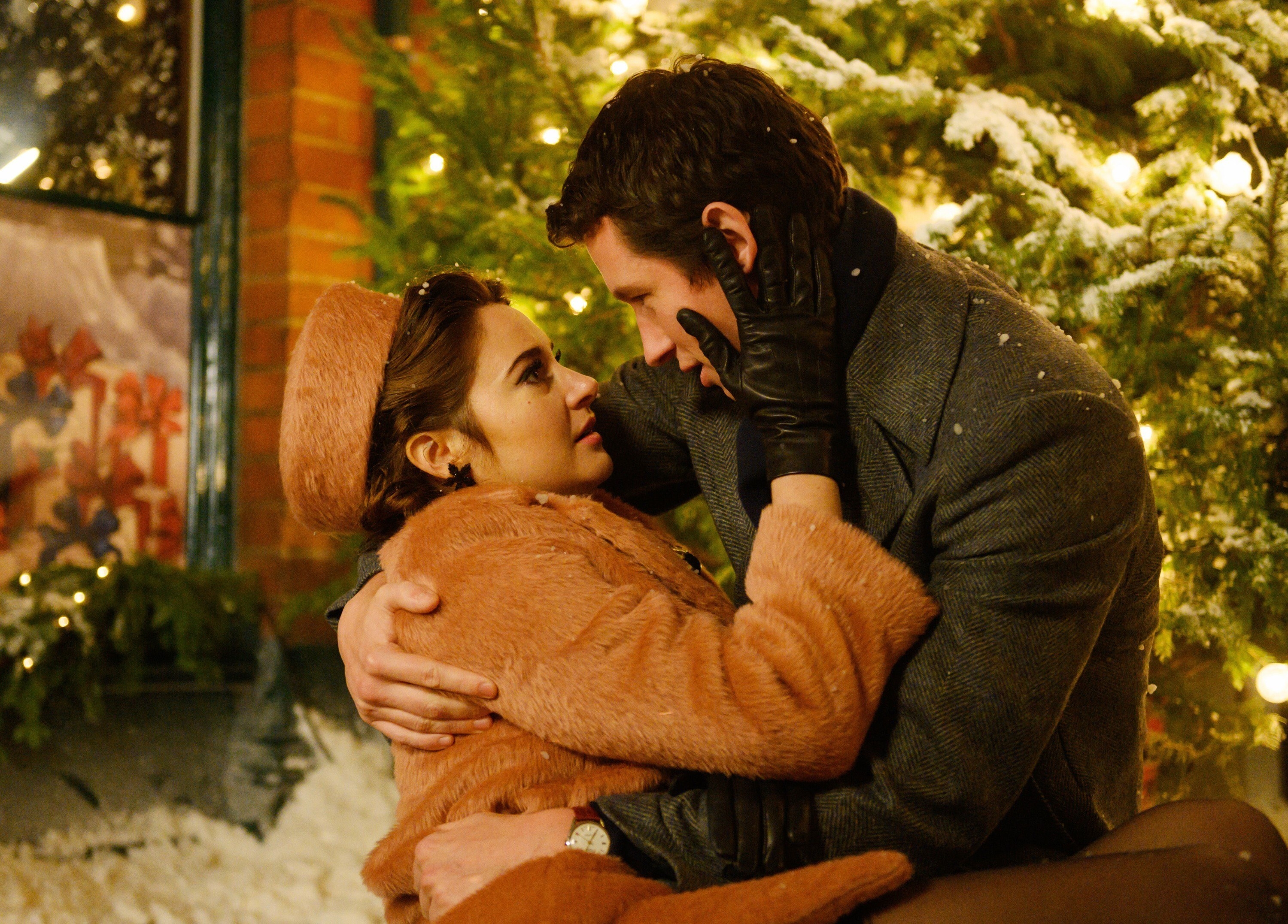 Anthony in wool overcoat leaning down to hold Jennifer, who has fallen on the snowy ground, wearing fuzzy coat and pillbox hat, gloves on, Christmas tree with lights behind them