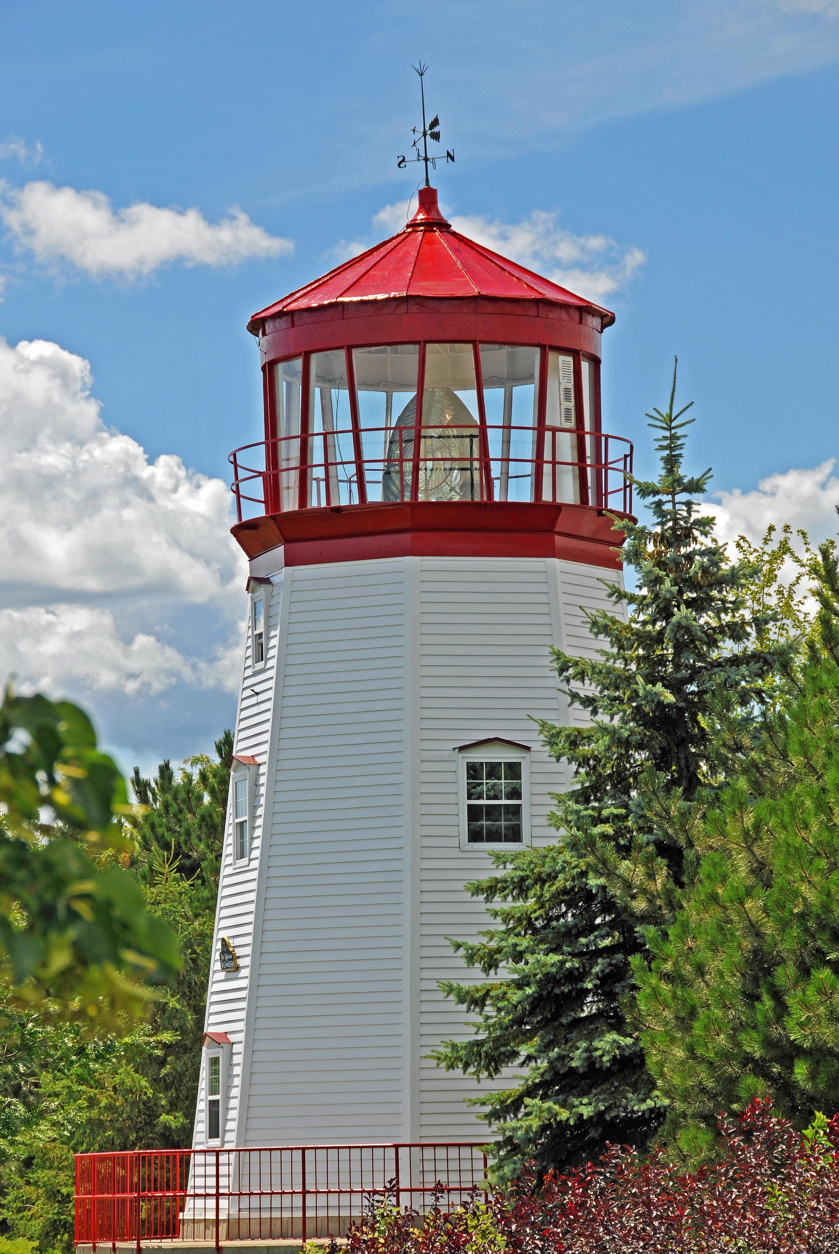 Photograph of a large white lighthouse with a red top, surrounded by greenery and a blue sky.