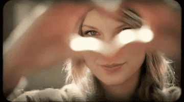 Taylor Swift making a heart with her hands then pointing towards the lens.