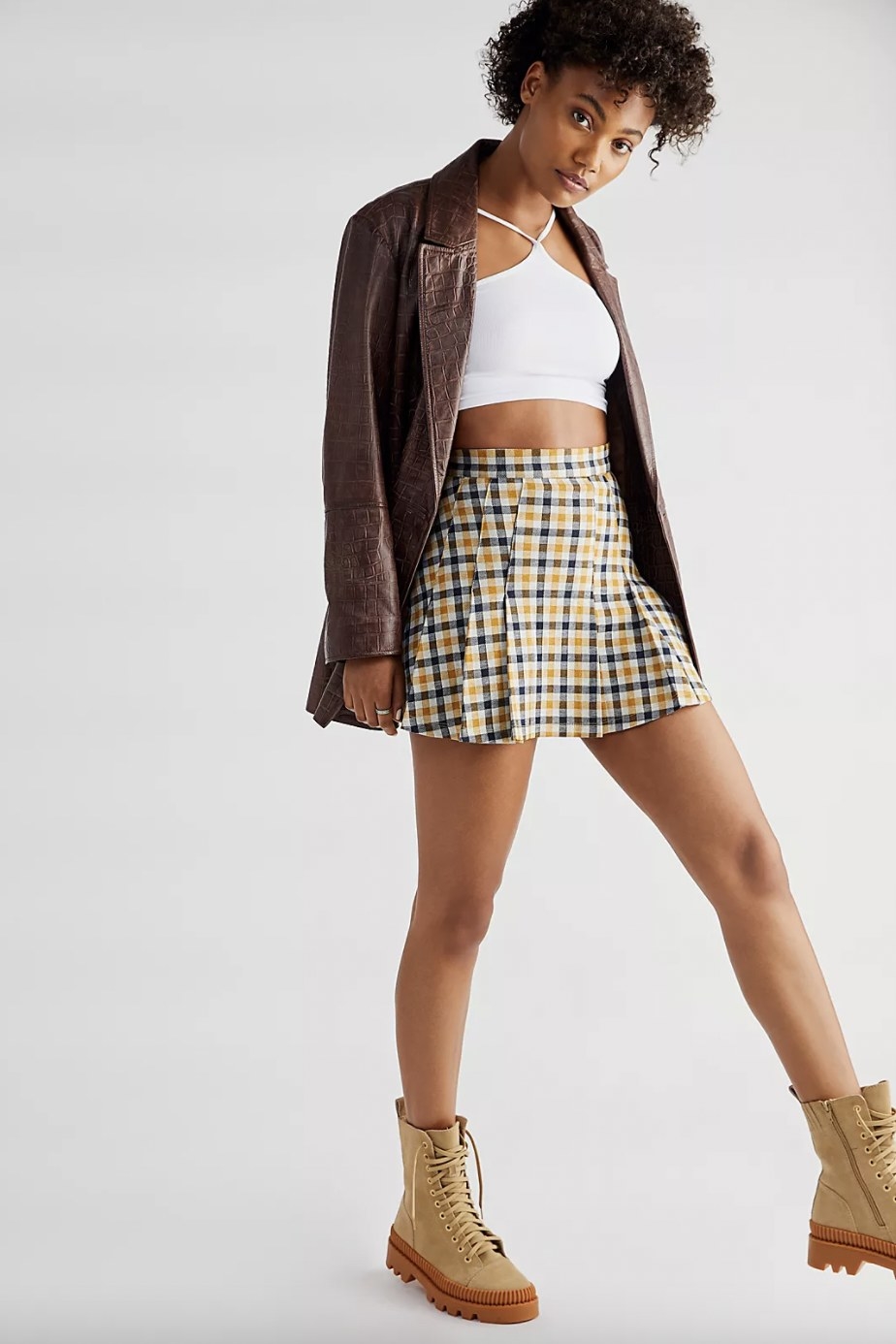 A woman wearing a brown jacket, a white shirt, a gingham print skirt and brown boots