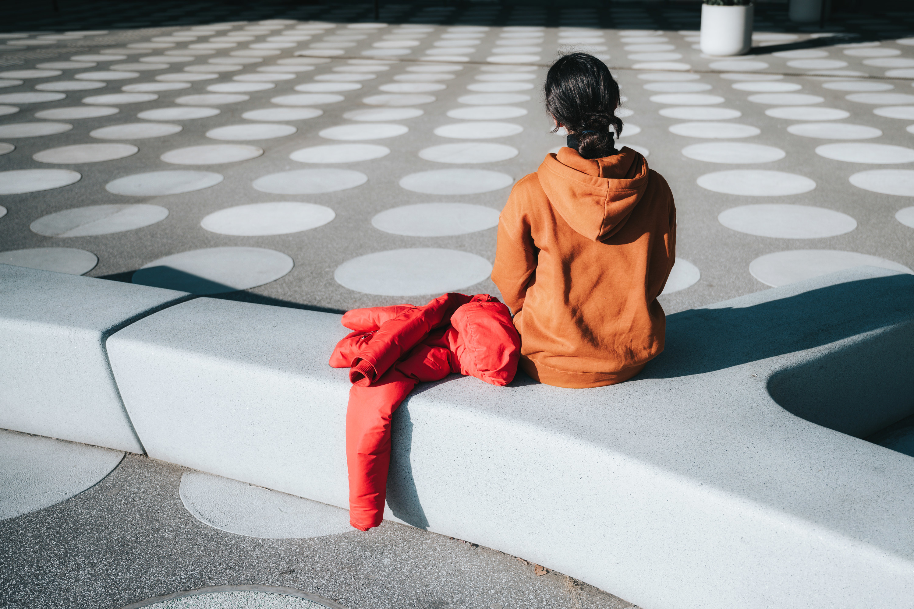 A woman in an orange hoodie sits on a bench in a public square