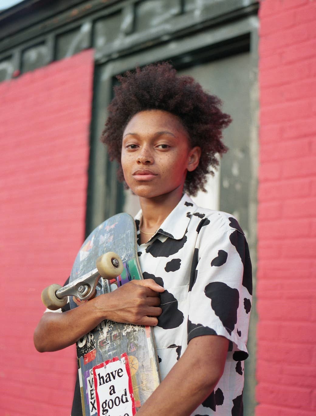 A young girl in a cow-print shirt holding a skateboard in front of a painted brick wall