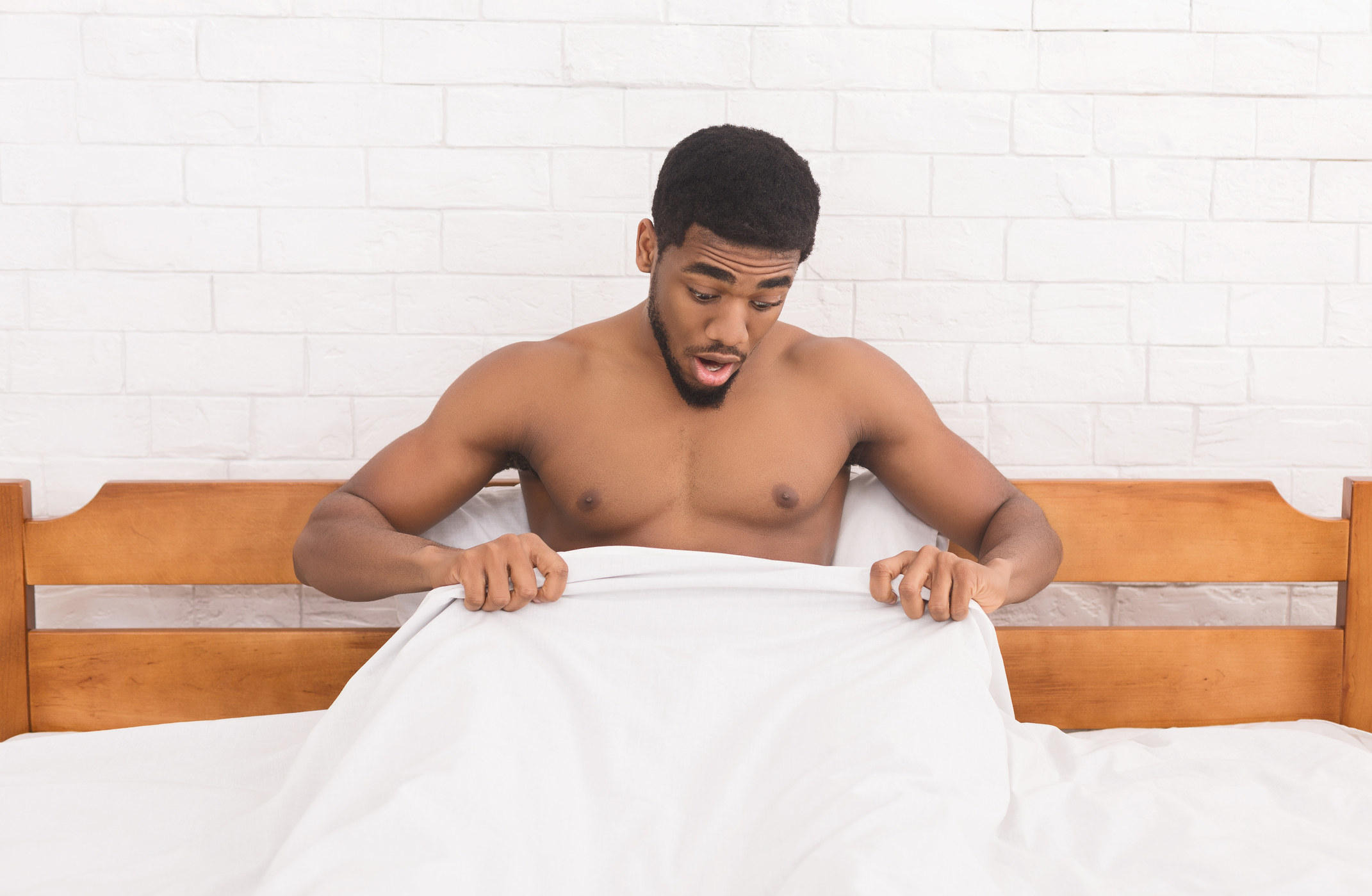 Man sitting up in bed and looking underneath the sheets at the bottom half of his body