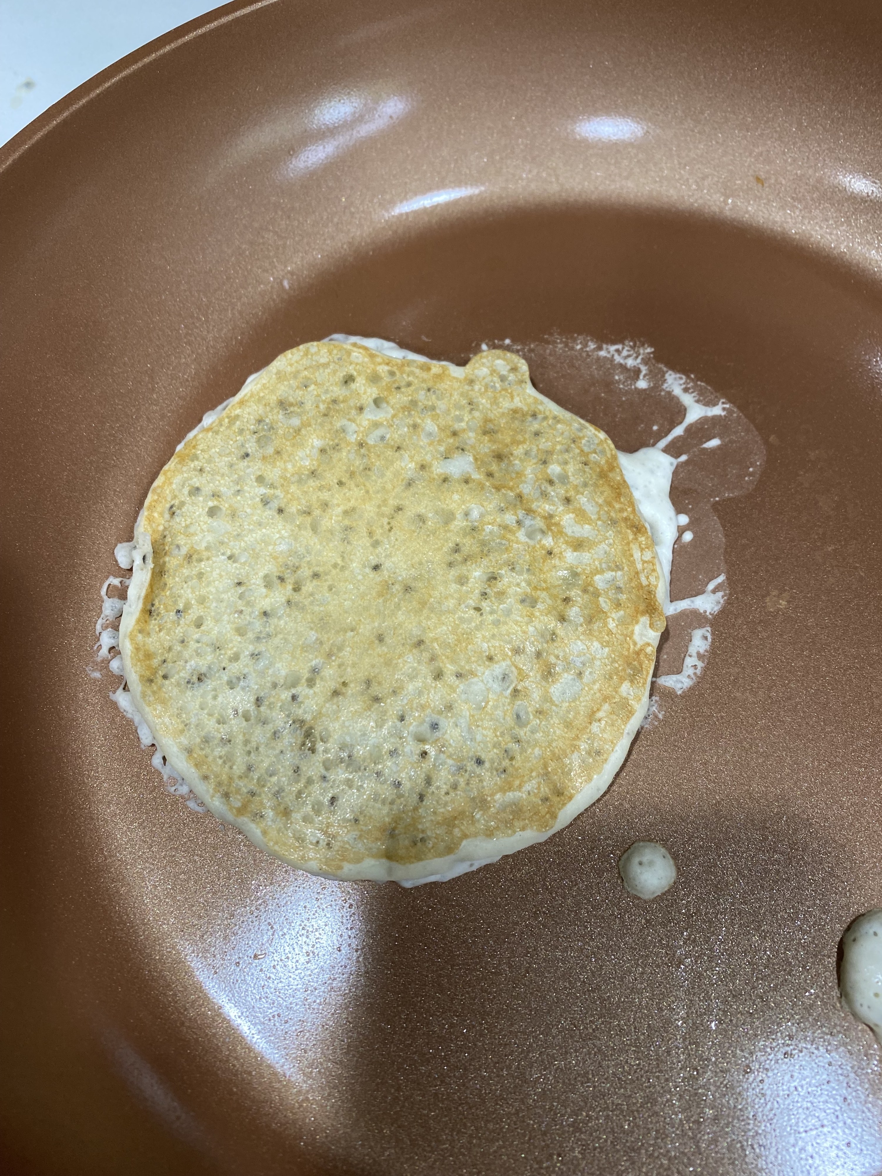 The pancake sizzling in the pan, with chia seeds dotted throughout the pancake