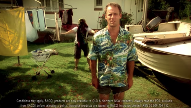 A man standing in his backyard in front of a boat and the clothes line; his wife is in the background playing a joke on him with a towel over her head