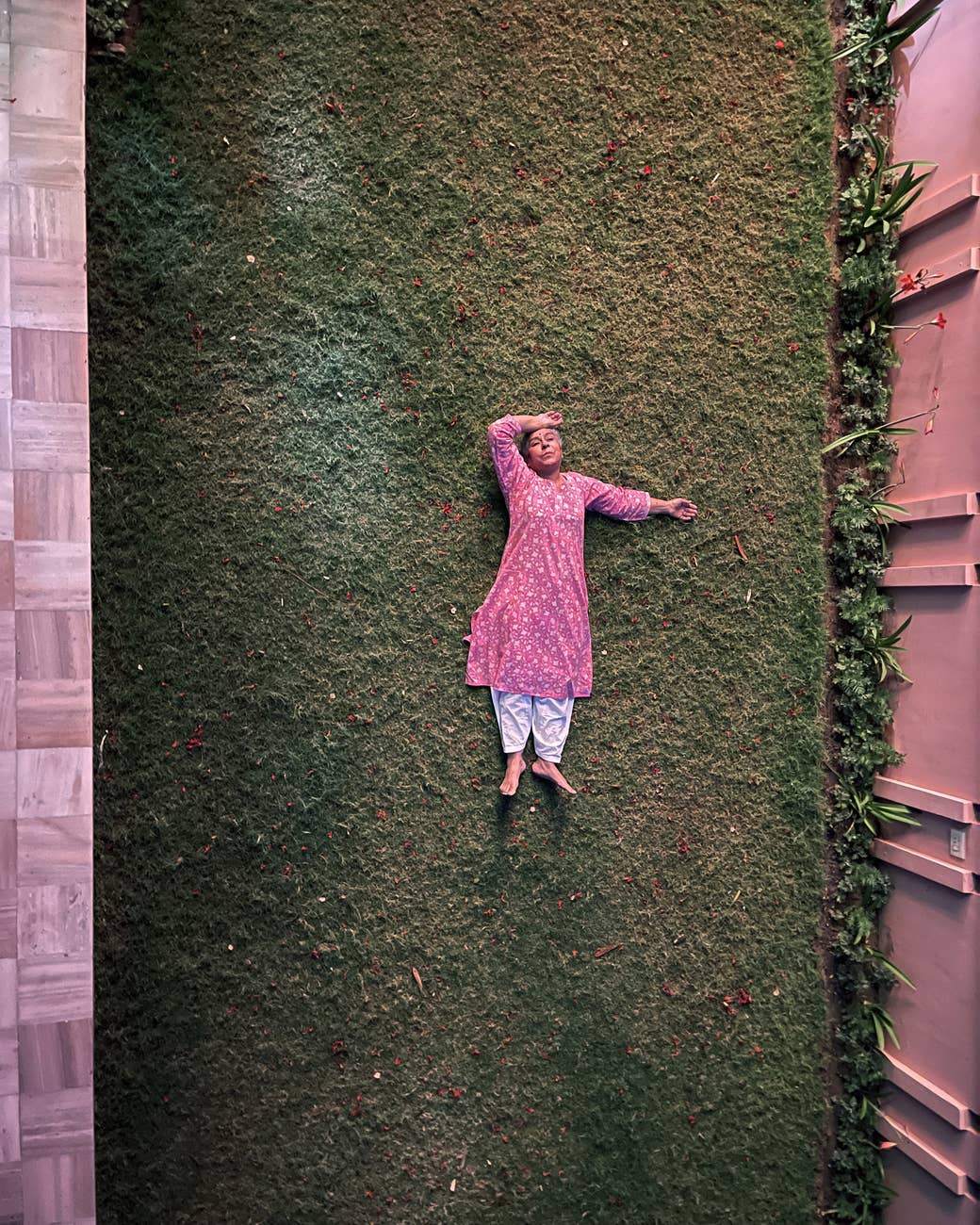 A woman lying on the ground in a garden with a hand to her head, seemingly at rest