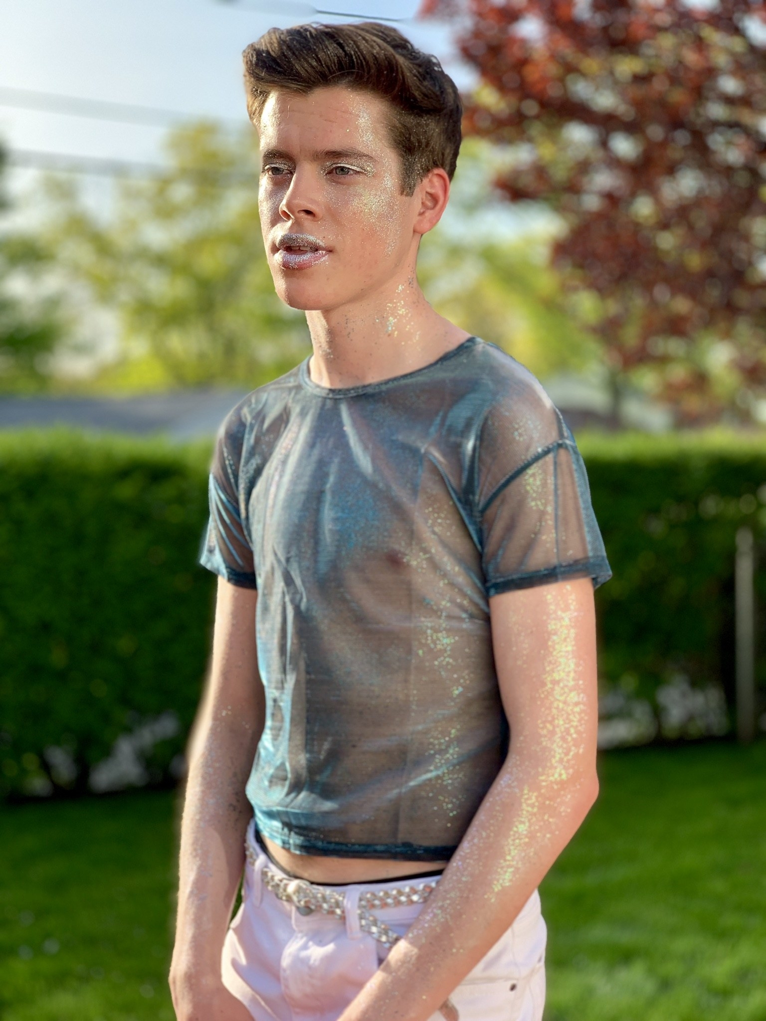 A young man covered in glitter and wearing a see-through T-shirt standing in a yard