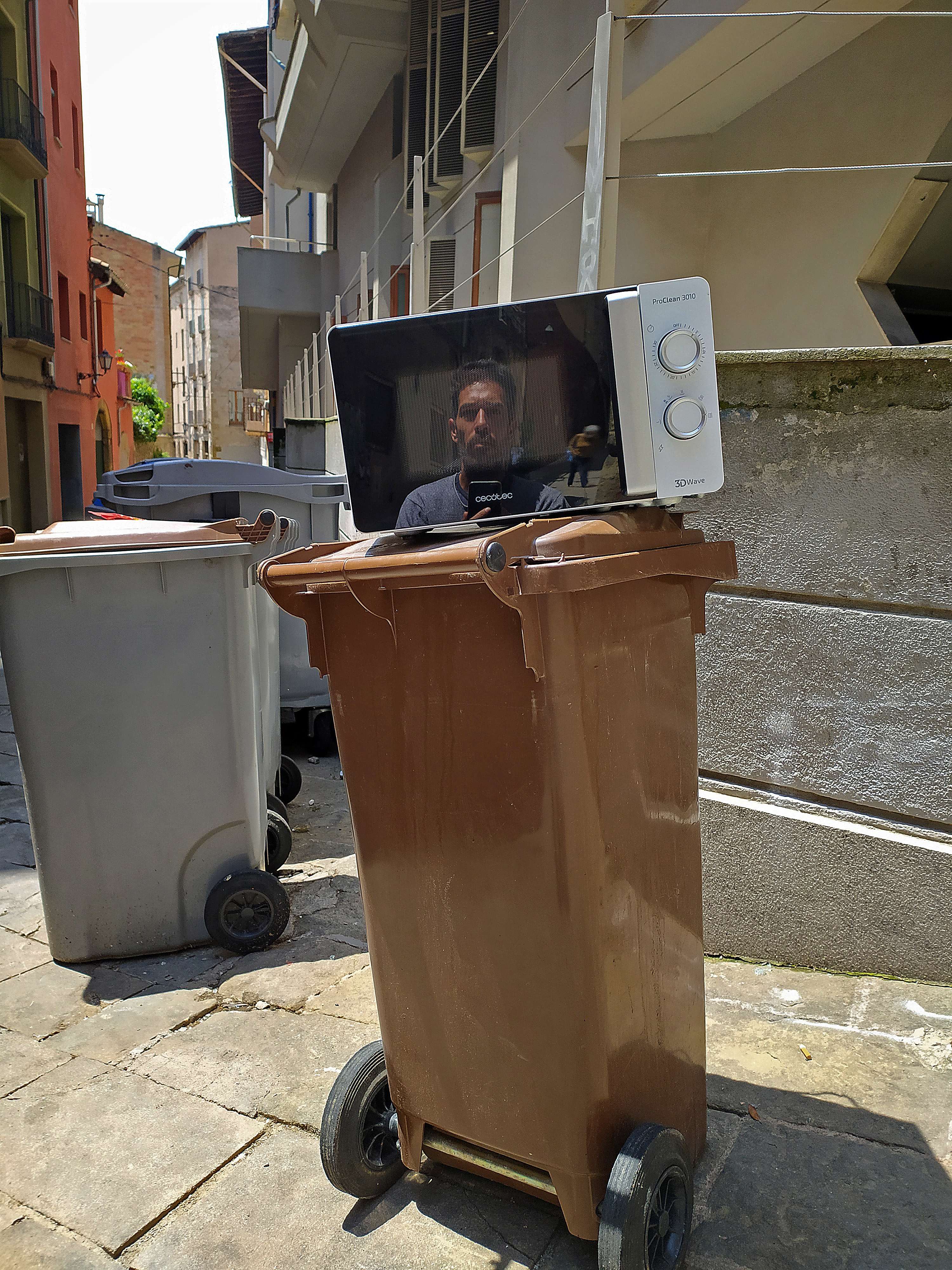 Someone is reflected in a microwave atop a garbage can on the street