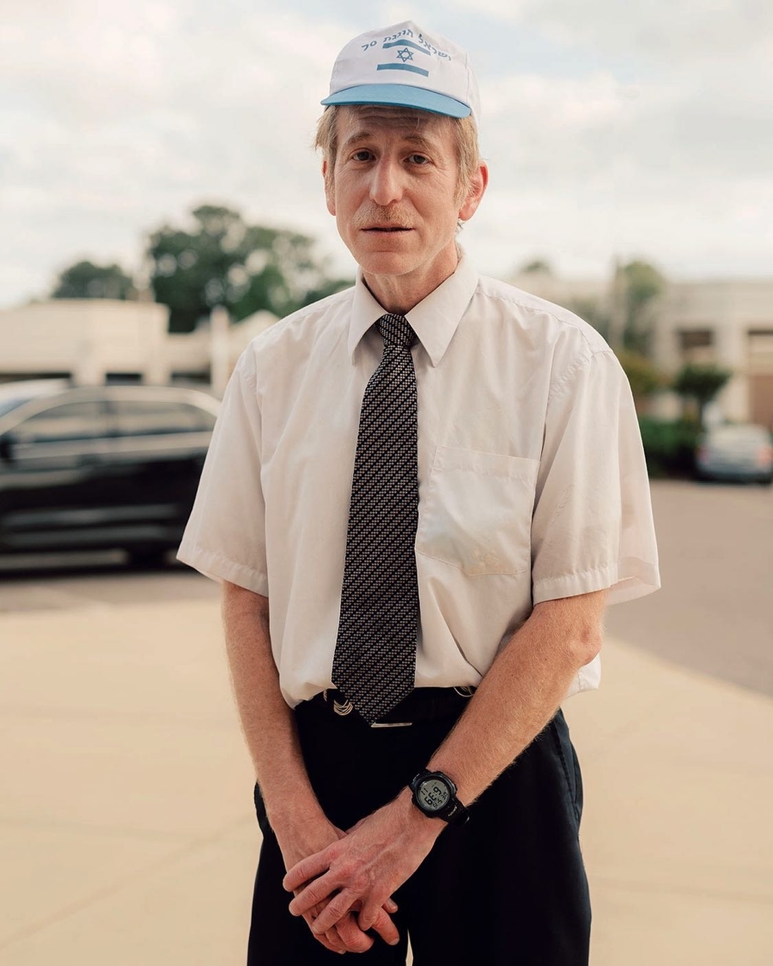 A man in a shirt and tie with a cap with the Israeli flag on it stands in a parking lot