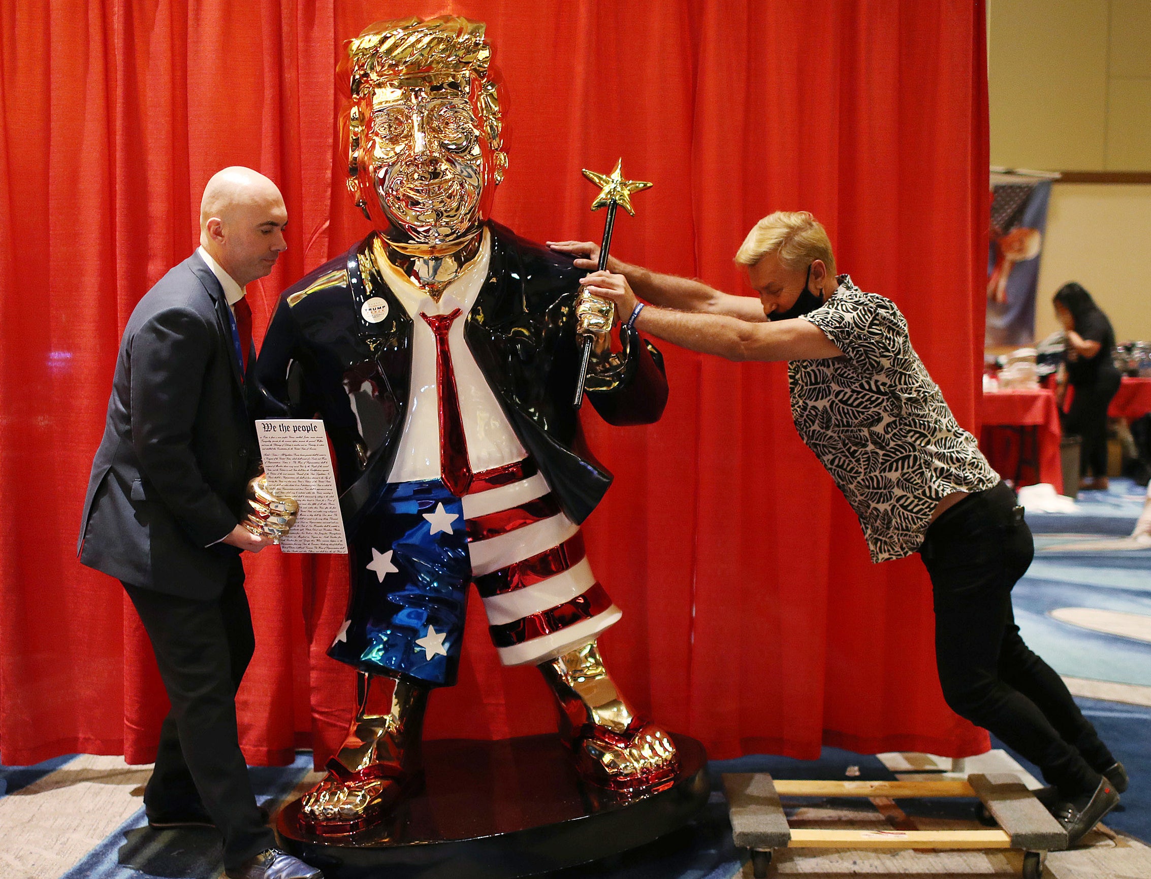 Two men stand on either side of a gold-plated statue resembling Trump, wearing American flag shorts, a suit, and tie, and carrying a wand with a star on the end
