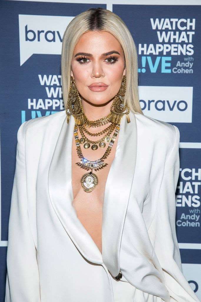 Khloé poses on a red carpet with straight hair while wearing plunging shirt jacket with chunky necklaces and earrings