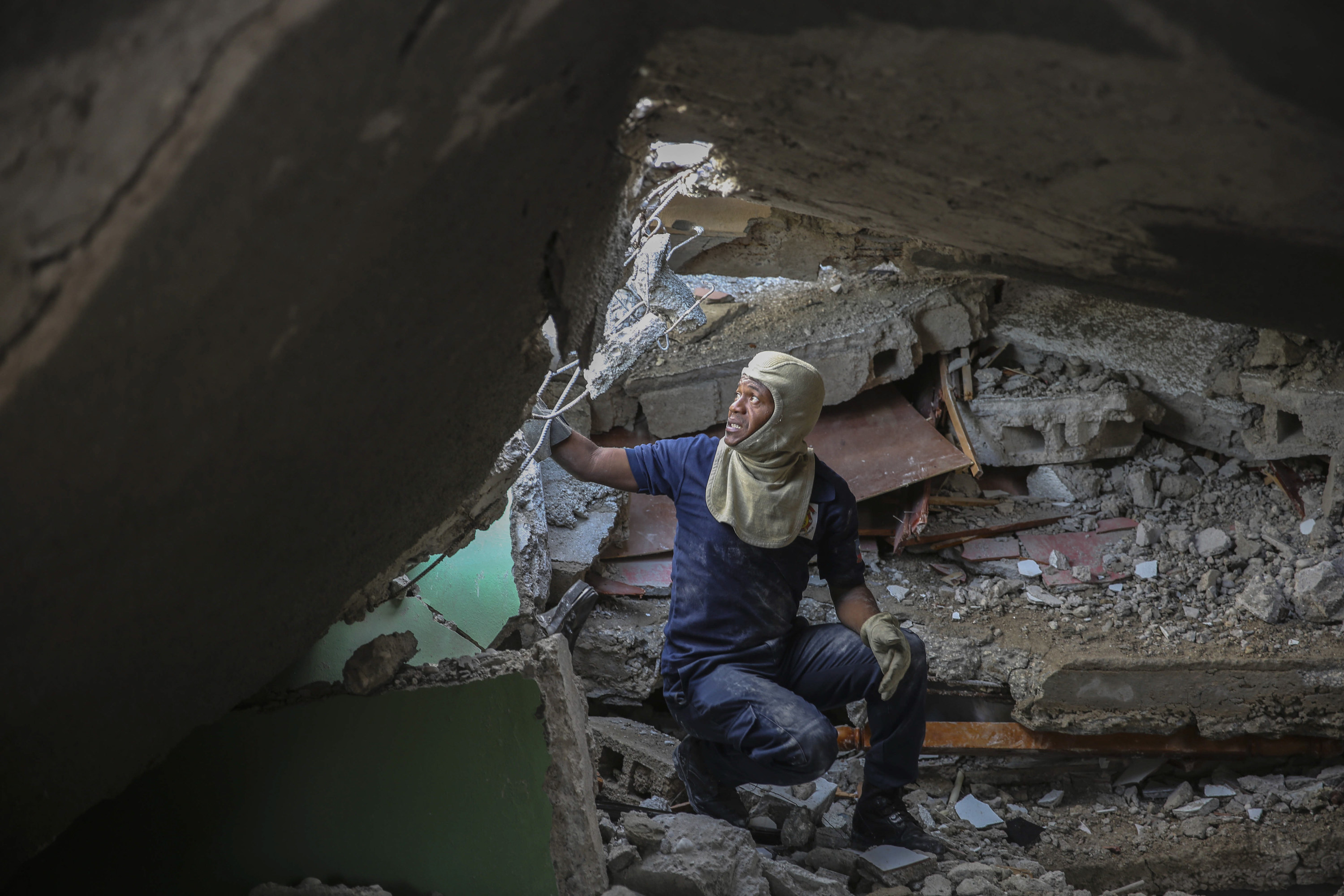 A man with protective headwear crouches and gazes upward amid rubble