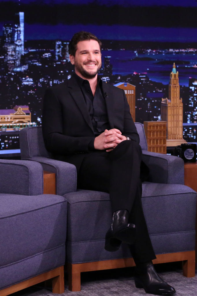 Kit smiling and sitting with his legs crossed and arms clasped