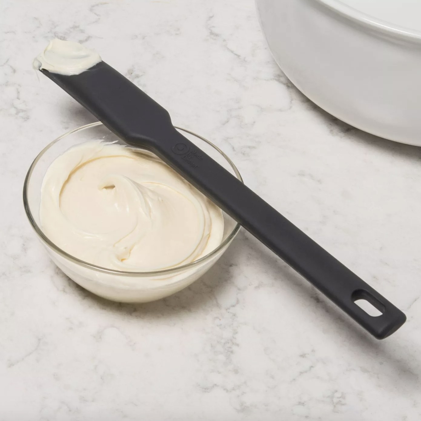The all black spatula is resting on top of a small glass dish of white cream texture with a bit on the edge