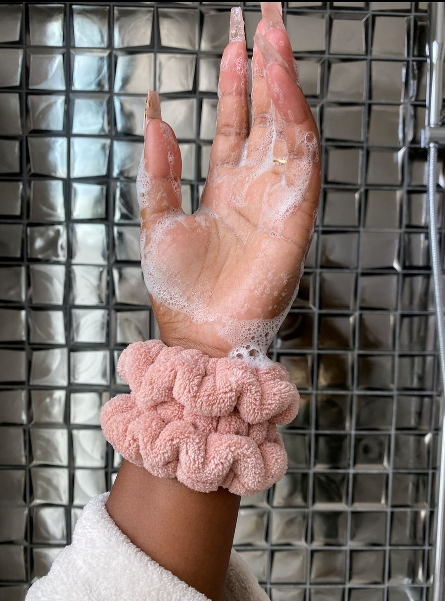 a hand wearing the cuff, which looks like a soft oversized scrunchie and is catching the suds from face wash, preventing it from running down the model&#x27;s arm