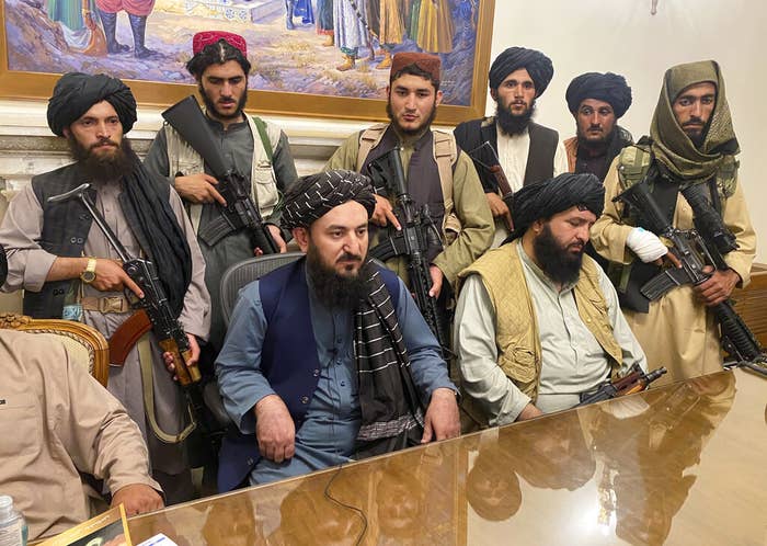 A group of men in headwear, most of them armed, sit at a table looking very solemn