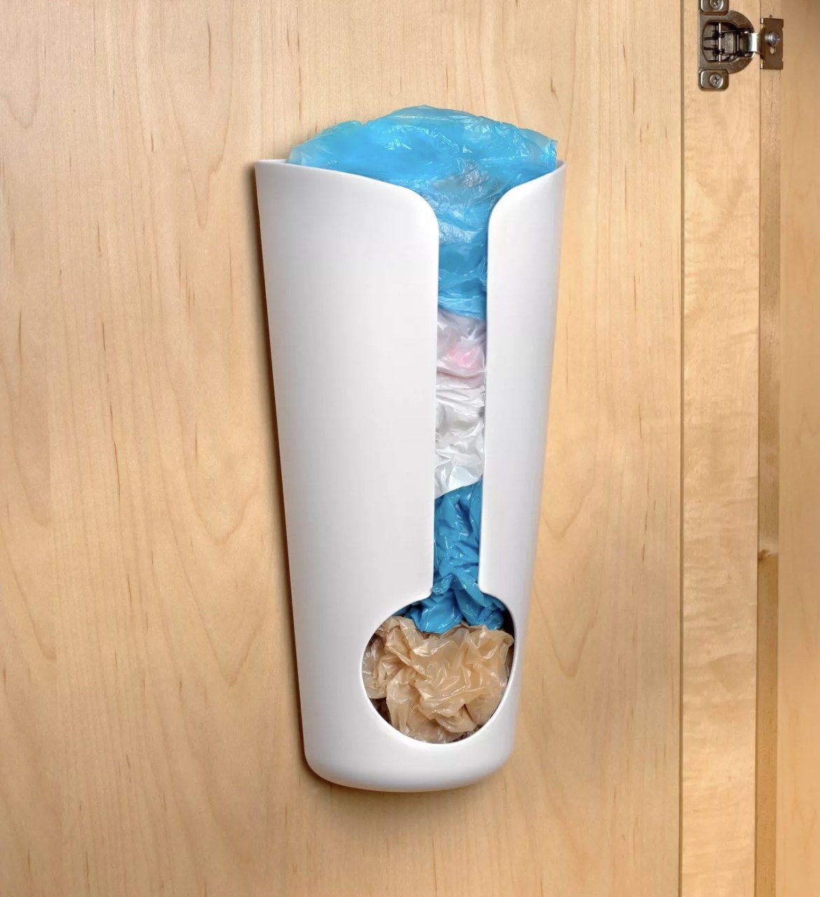 The white plastic bag holder is attached to a light wooden cabinet and structurally has a thin rectangular slot that widens into a spherical shape and is holding various colored plastic bags