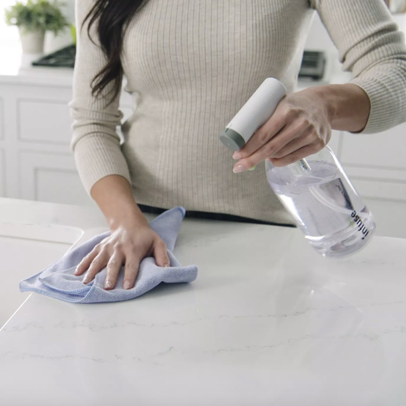 An adult is using the clear spray bottle that says &quot;infuse&quot; and has a white nozzle with a grey circular edge to wipe down a marble countertop