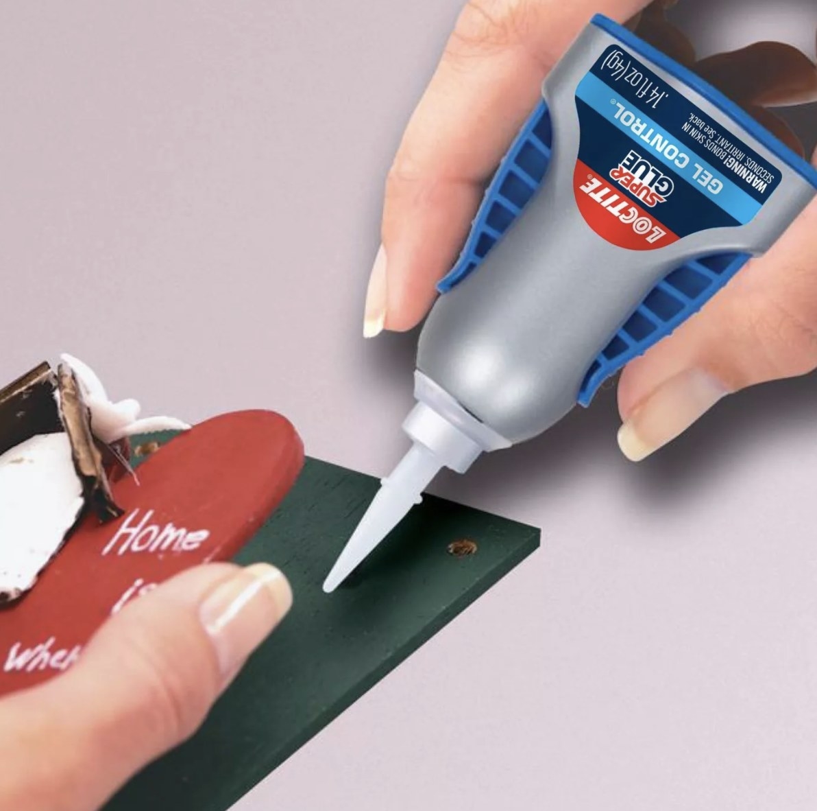 The grey and blue bottle of glue that says &quot;LOCTITE&quot; &quot;SUPER GLUE&quot; and &quot;GEL CONTROL&quot; is gluing a green and red home decoration back together that has the words &quot;Home is where&quot; visible