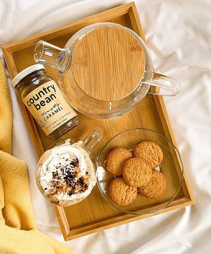 The jar of coffee place on a wooden tray with a mug of frothy coffee, plate of cookies, and kettle