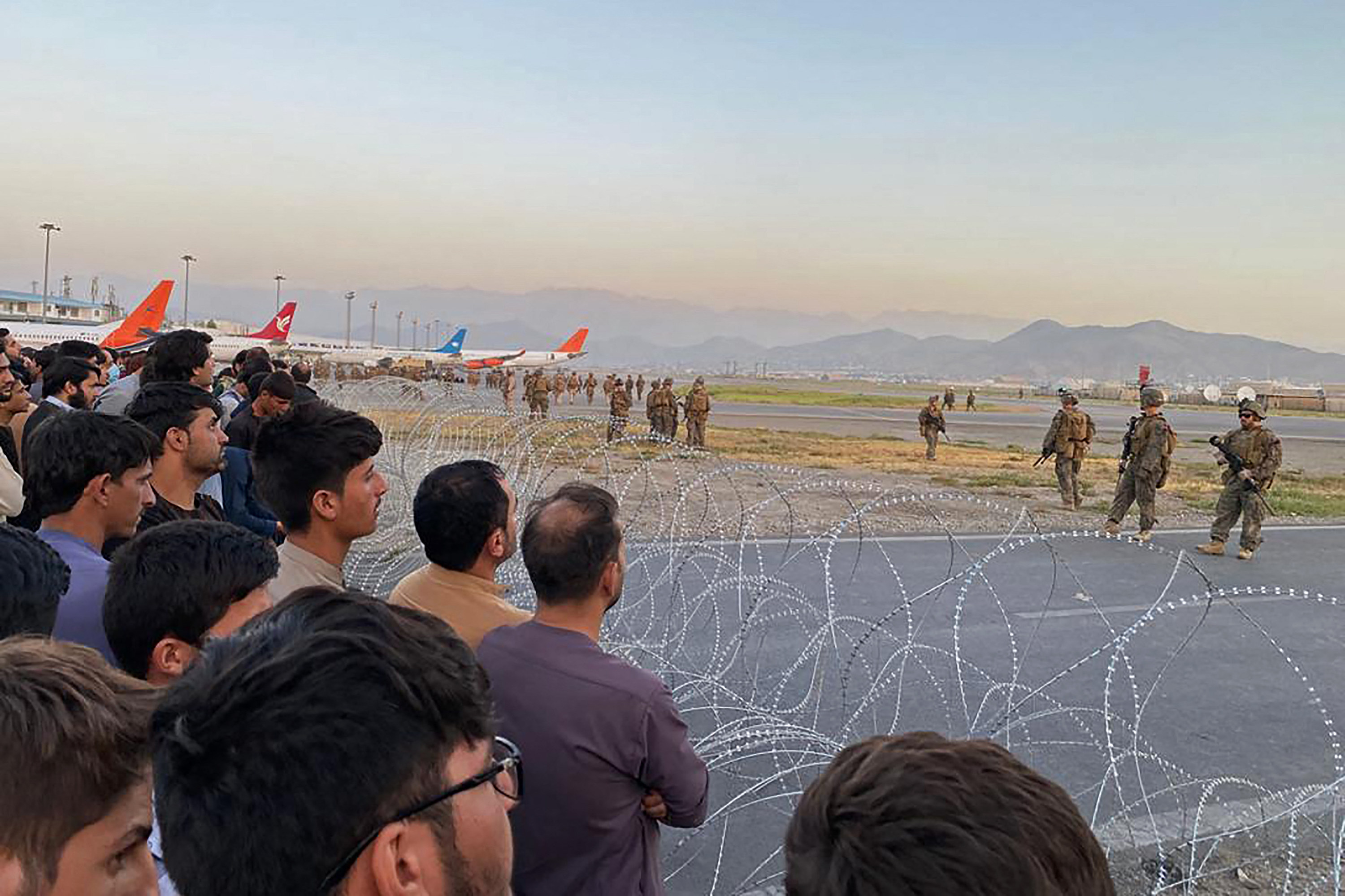 People behind barbed wire look out at planes and soldiers at the airport