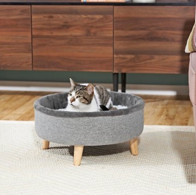 A cat lying in a grey, elevated round bed on four wooden legs